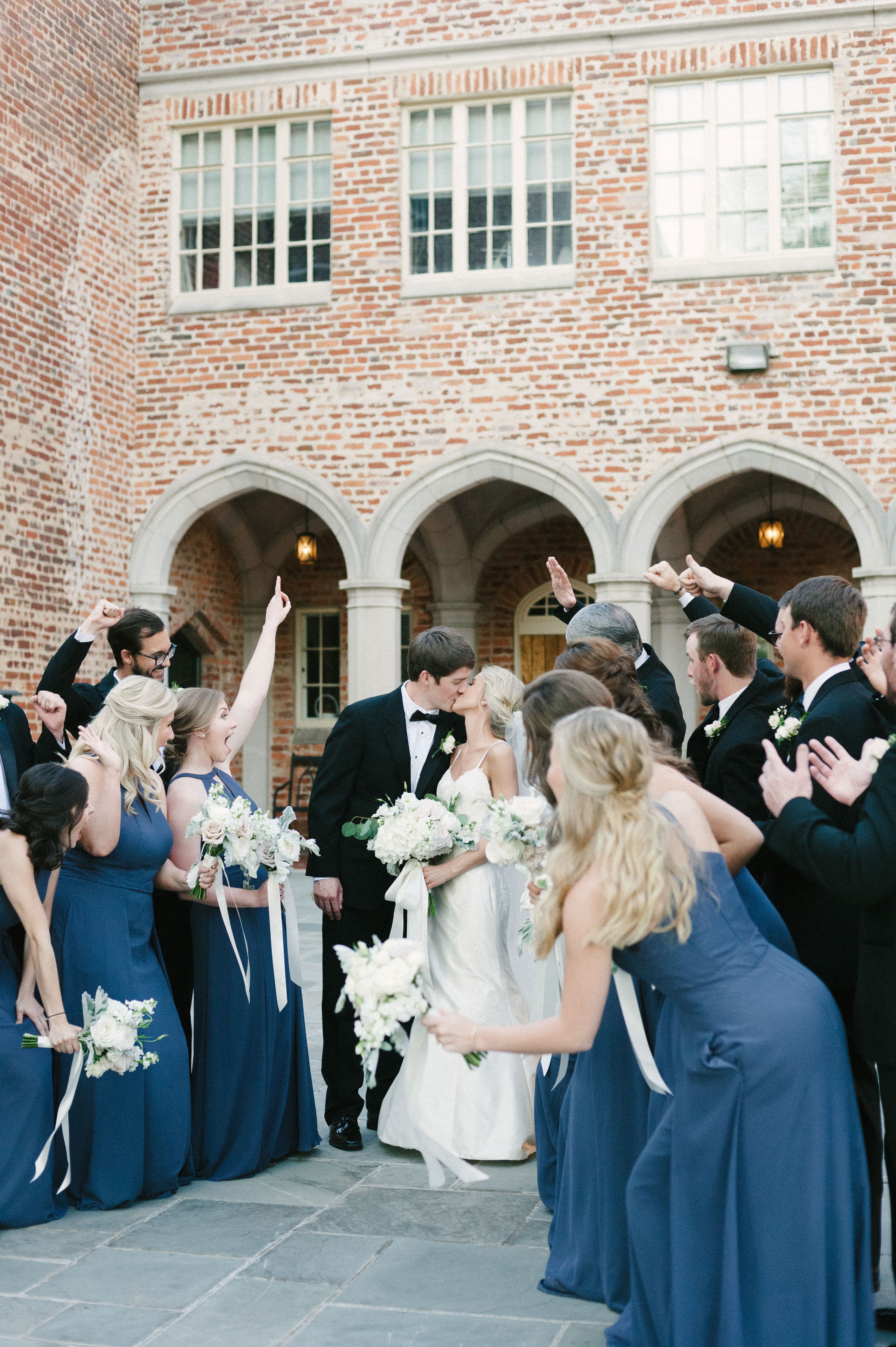 The ultimate wedding party getting excited as a newly married couple kisses in front of them in the courtyard at First Baptist Church in Selma, AL by Olivia Joy Photography