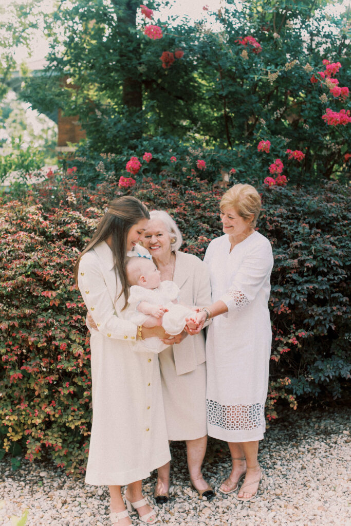A portrait of four generations happy and smiling at the youngest in their white outfits in front of a flowering bush