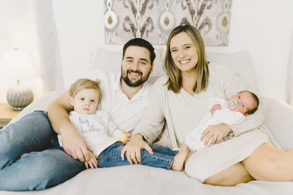 Happy family of four with almost 2 year old and newborn at their house on their bed smiling at the camera