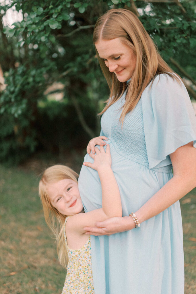 Maternity photos. A little girl hugging her pregnant mommy's belly while she looks and smiles at the camera and her mom looking at her 